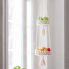 White macramé plant hanger hanging by the window. There are fruits in the baskets of the layered hanger.