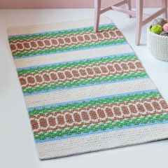 Free pattern: Anja Rug designed by Molla Mills on the floor next to a chair and a yarn basket. The rug is crocheted with Lankava Essi Tube Yarn using colours natural white 2, light blue 18, green 10 and light brown 4.